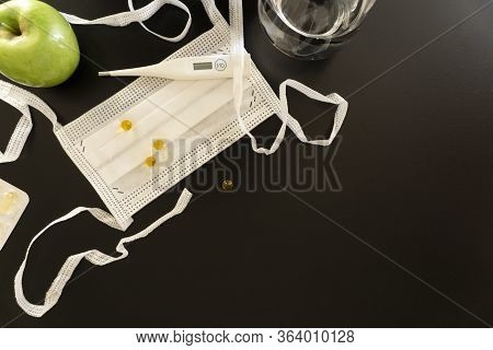 A Medical White Protective Mask, A White Thermometer, Several Vitamin Pills And A Green Apple Lie On