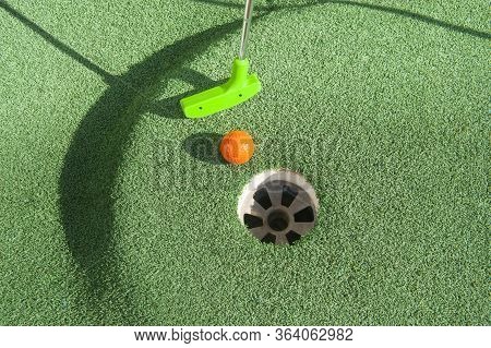 Green Mini Golf Club Puts And Orange Ball Towards A Hole On Green Turf
