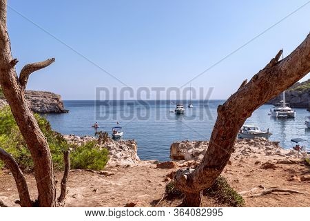 Seascape View Of The Famous Bay Cala Turqueta With Trees In The Foreground And Boats In The Sea. Men