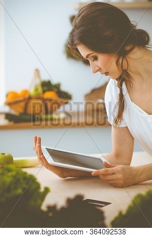 Young Woman Looking For A New Recipe For Cooking In A Kitchen. Housewife Is Making Online Shopping B