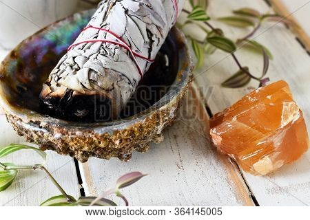 A Close Up Image Of A White Sage Bundle In An Abalone Shell With Honey Calcite On A White Wooden Tab