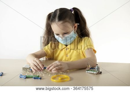 A Girl In A Protective Mask At The Table Plays A Constructor. Home Insulation. World Pandemic.