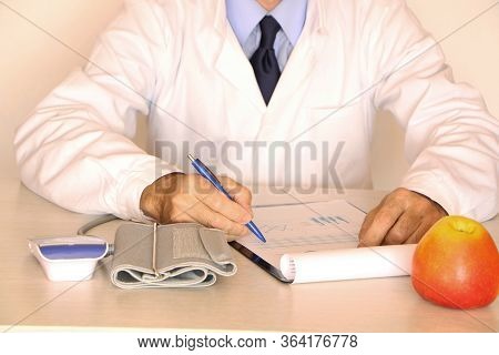 Close-up - The Doctor In A White Coat Sits At The Table And Eats A Healthy Apple.