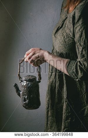 Woman In Dark Dress Hold In Hands Craft Handmade Ceramic Teapot With Twig Wooden Handle For Serving 