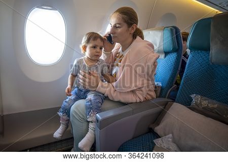 A Young Caucasian Mother Sits In An Airplane Chair And Talks On The Phone, In Her Arms Sits A Little