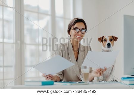 Positive Female Entrepreneur Poses At Workplace, Holds Paper Documents, Sits In Front Of Computer, W
