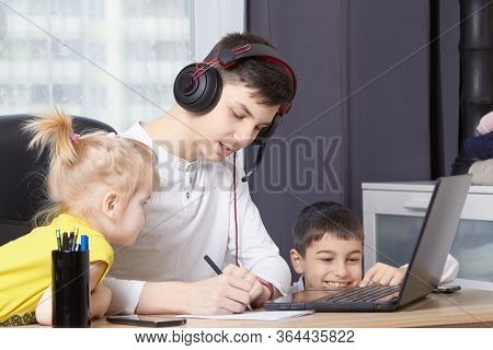 Teenager Looks At A Laptop And Writes Down A Task And Children Interfere With Learning