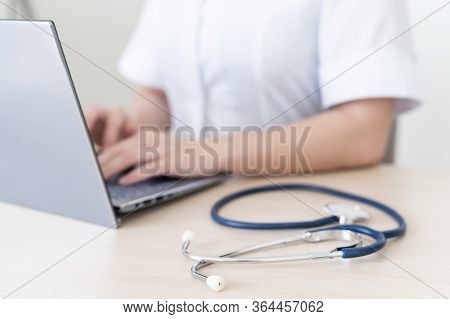 Unrecognizable Female Doctor In Uniform Uses A Laptop. Faceless Medical Worker Writes A Report On Th