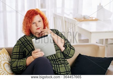 Pensive Young Pluz Size Woman Sitting On Sofa And Reading Article On Tablet Computer