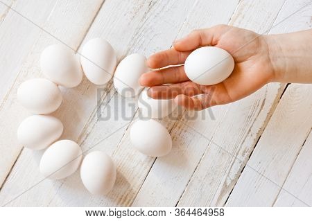 A White Chicken Egg Lying In A Man’s Hand, Shot On A White Painted Wooden Surface. Background For Li