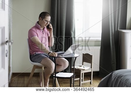 The Man Is Siiting By The Small Table And Working At Home On Laptop With The Phone. Man In Quarantin