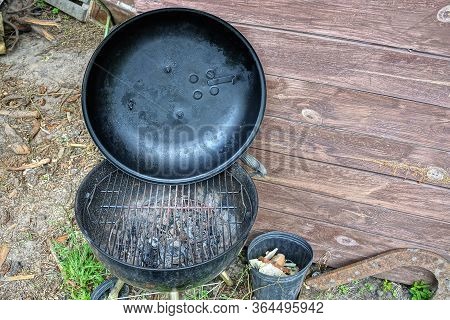 Round Iron Black Barbecue With An Open Lid And A Metal Grill And Ash On The Street Against A Brown W