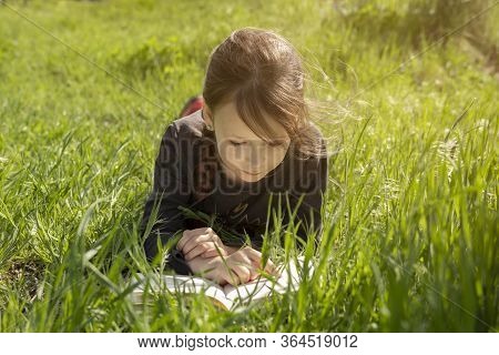 A Little Girl Is Lying On Her Stomach Reading On The Grass. She Has A Look Of Enjoyment On Her Face 