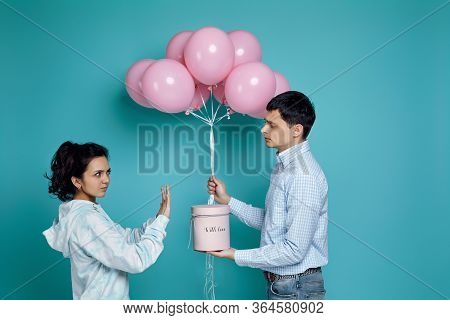 Young Man Presenting Gift Box And Pink Air Balloons To His Upset Girlfried On Blue Background. Woman