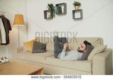 Restful man in casualwear lying on couch with his head on pillows in front of tv set and watching movie or broadcast in living-room