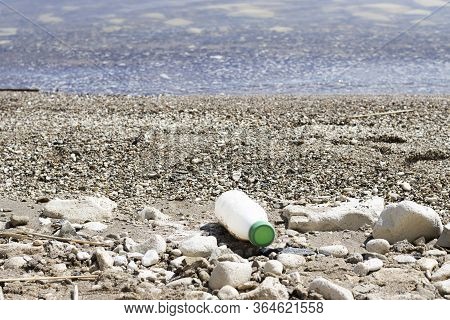 White Plastic Bottle With A Green Lid On The Bank Of The River, Sea. Concept Of Pollution Of Rivers,
