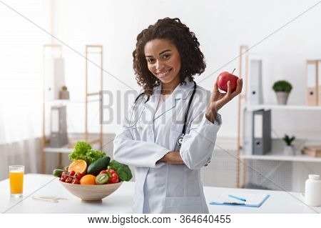 Positive Black Woman Nutritionist Posing At Clinic, Holding Apple, Copy Space