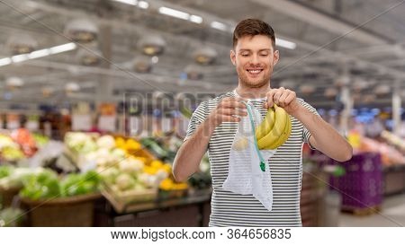 food, sustainability and eco living concept - smiling young man in striped t-shirt putting bananas into reusable string bag over market or grocery store on background