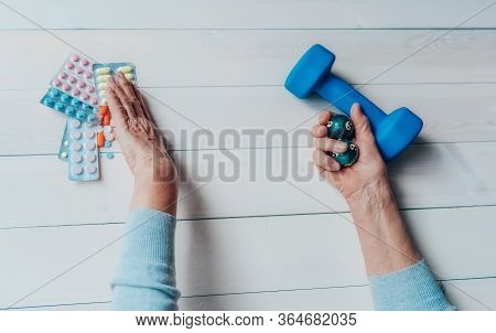 Senior Wrinkled Hand Pushing Back Pills And Thermometer, Dumb-bell On A Table. Another Old Woman Han