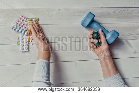 Senior Wrinkled Hand Pushing Back Pills And Thermometer, Dumb-bell On A Table. Another Old Woman Han