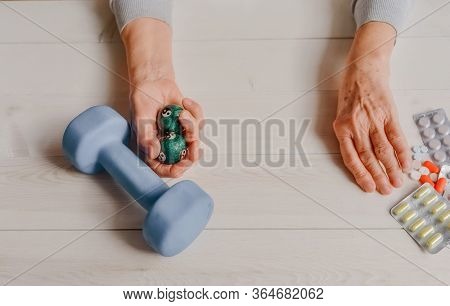 Senior Wrinkled Hand Pushing Back Pills And Thermometer, Dumb-bell On A Table. Another Old Woman Han
