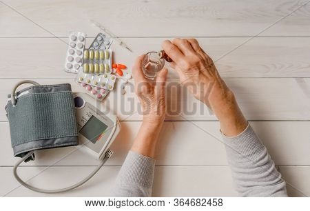 Senior Hands With Medicine Bottle Putting Drops In A Glass, Pills, Tonometer, Thermometer On A Table