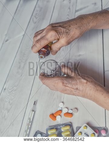 Senior Hands With Medicine Bottle Putting Drops In A Glass, Pills, Tonometer, Thermometer On A Table