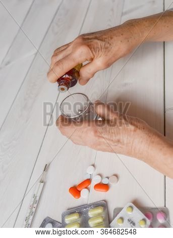 Senior Hands With Medicine Bottle Putting Drops In A Glass, Pills, Tonometer, Thermometer On A Table