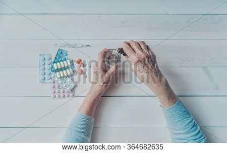 Senior Hands With Medicine Bottle Putting Drops In A Glass, Pills, Tonometer, Thermometer On A Table