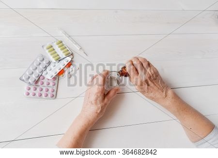 Senior Hands With Medicine Bottle Putting Drops In A Glass, Pills, Tonometer, Thermometer On A Table