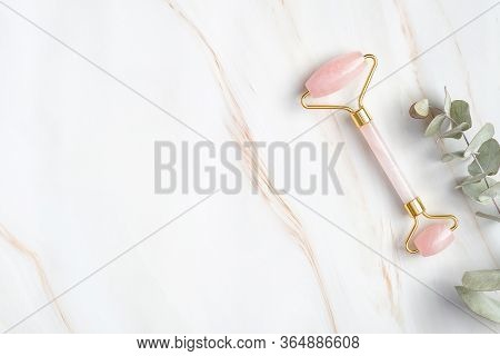 Rose Quartz Face Roller And Eucalyptus Leaf On Marble Background, Top View. Face Massage Tool, Beaut