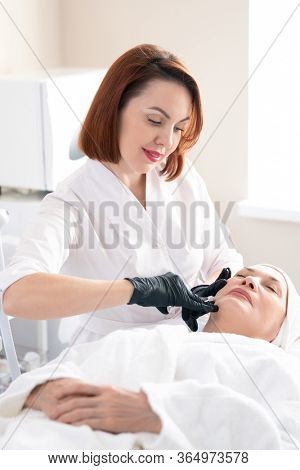 Smiling redhead beautician in white coat making skin-boosting injection to mature woman at beauty treatment procedure