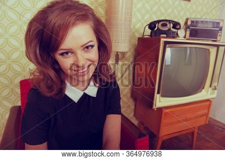 Young smiling ecstatic woman looking at camera, smiling in room with vintage wallpaper and interior, retro stylization 60-70s. Furniture, tv set and another technique of the mid 20th century