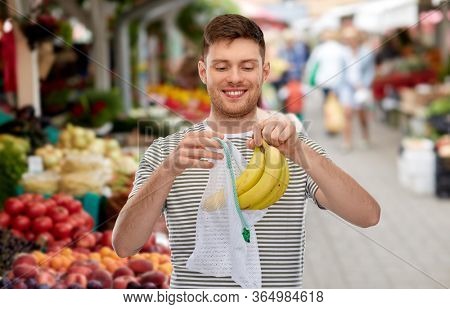 food, sustainability and eco living concept - smiling young man in striped t-shirt putting bananas into reusable string bag over street market on background