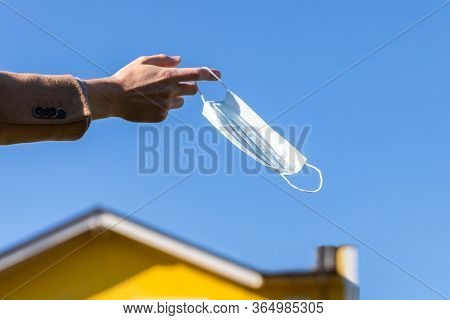 Man Takes Off Medical Protective Mask And Holds It On Her Finger On Blue Sky And Yellow House On Bac