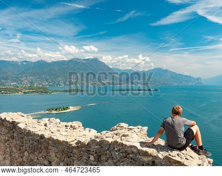 Boy Sit  Alone On Rest Of Manerba Castle Wall Above Lago Di Garda Lake. Summer Holiday And Travel, O