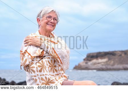 Portrait Of Smiling Senior Woman Wearing Eyeglasses And Scarf Sitting Outdoors At Sea Enjoying Good 