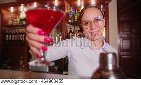 Female Bartender In Eyeglasses With Cocktail Standing Behind Bar, Looking At Camera, Offering Alcoho