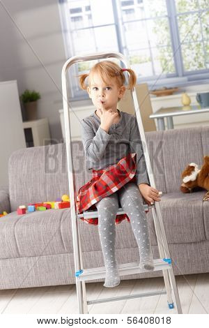 Cute little girl sitting impishly on ladder, putting forefinger on lips as calling for silence.
