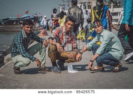 MUMBAI, INDIA - 09 JANUARY 2015: Three Indian boys crouch next to sea while people pass by. Post-processed with grain, texture and colour effect.