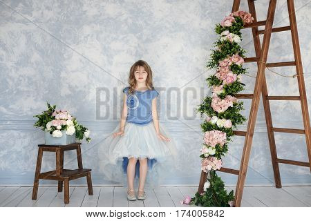 Beautiful and charming girl standing in the studio in a beautiful blue dress blue background and decor as stairs and flowers