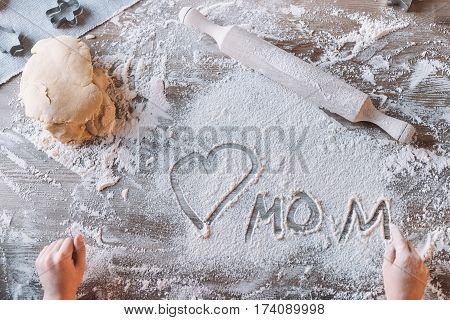 'Partial top view of child drawing heart symbol and word mom in flour on table Mothers day concept