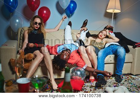 Two beautiful girls relaxing on sofa after New Year party while their drunken male friends having nap, all floor covered with confetti