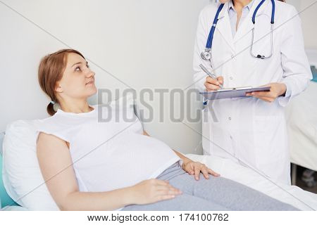 Young pregnant patient lying in hospital bed while doctor asking her questions about her health