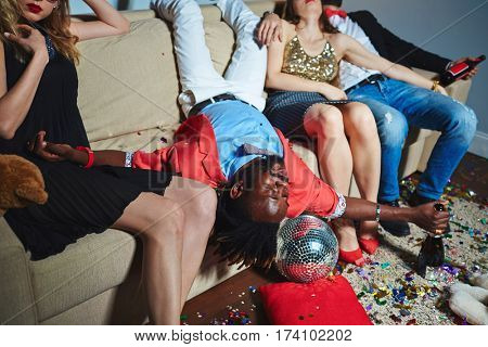 Portrait of middle-aged Afro-American man lying upside-down on couch while his friends sitting next to him with closed eyes after night party