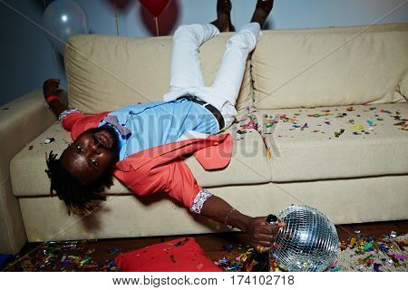 Tipsy African American man with short dreadlocks lying upside-down on brown sofa and looking at camera after New Year party