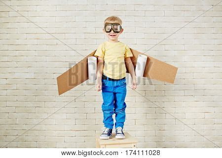 Funny little boy in aviator goggles and with carton airplane wings behind his back standing on wooden chair and looking at camera with smile