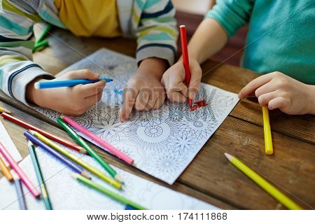 Close-up shot of two pairs of little hands coloring mandala with felt-tip pens, pencils of different colors and illustrations laid on wooden table