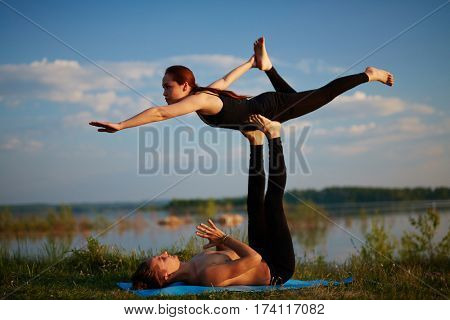 Sporty couple balancing in front bird pose while practicing acro yoga on river bank in summer