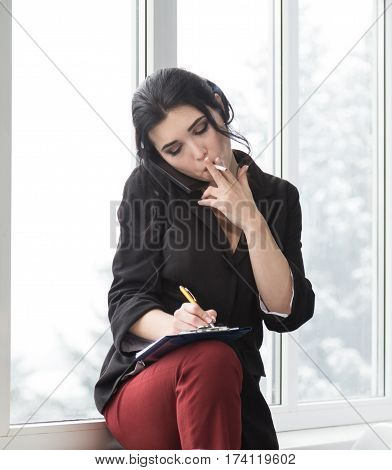 Business woman in office with a cigarette speaking on mobile phone and holding pen with notepad